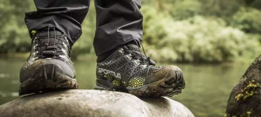A person in muddy hiking boots standing on a rock by a river