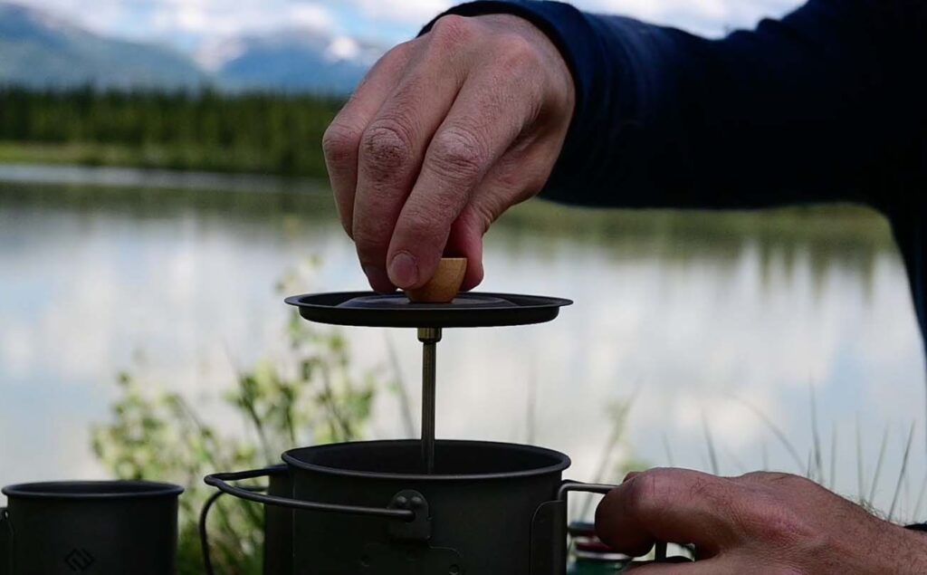 A hand pressing down a French press coffee maker outdoors by a lake