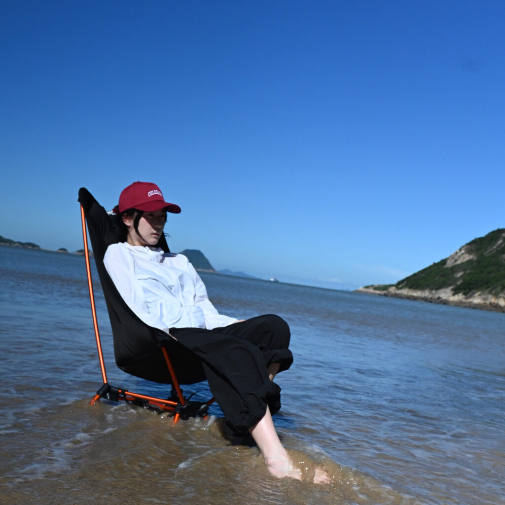 A girl in a white shirt and black pants with a red hat sits on a beach chair with their feet in the water, relaxing under a clear blue sky.