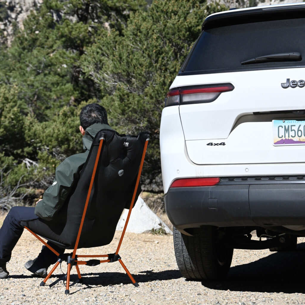Person sitting in a black camping chair with orange frame next to a white Jeep