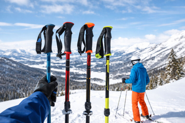 Four ski poles displayed on a snowy mountain viewpoint with a skier in the background, illustrating a beginner ski poles buying guide for 2026