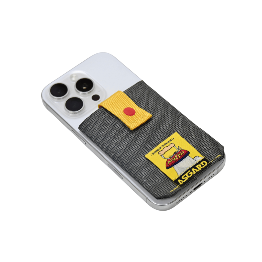 Dark gray Asgard card holder attached to the back of a silver phone, with its yellow strap and red snap visible.
