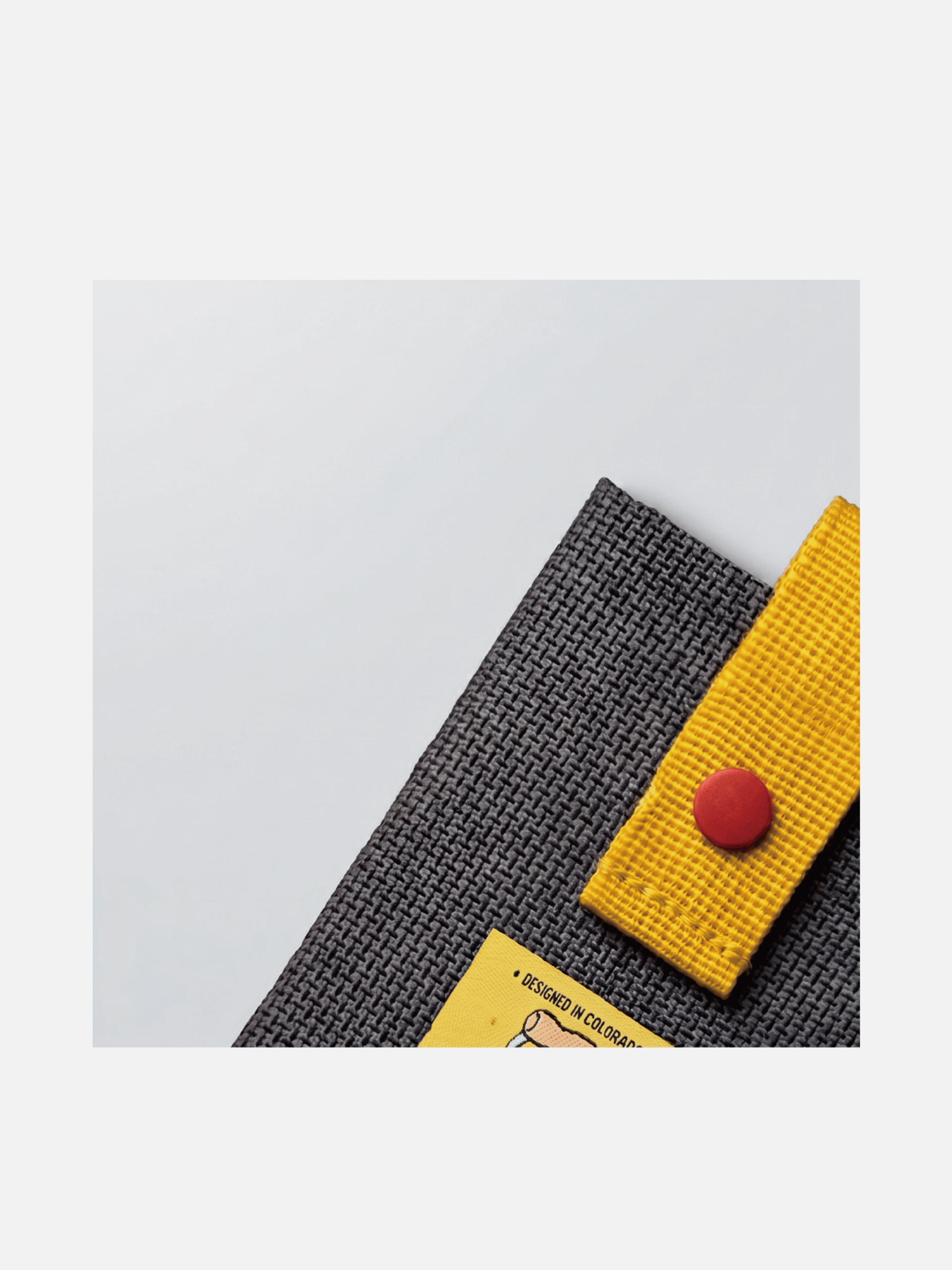Close-up of the card holder’s dark gray woven fabric, yellow strap and red snap.
