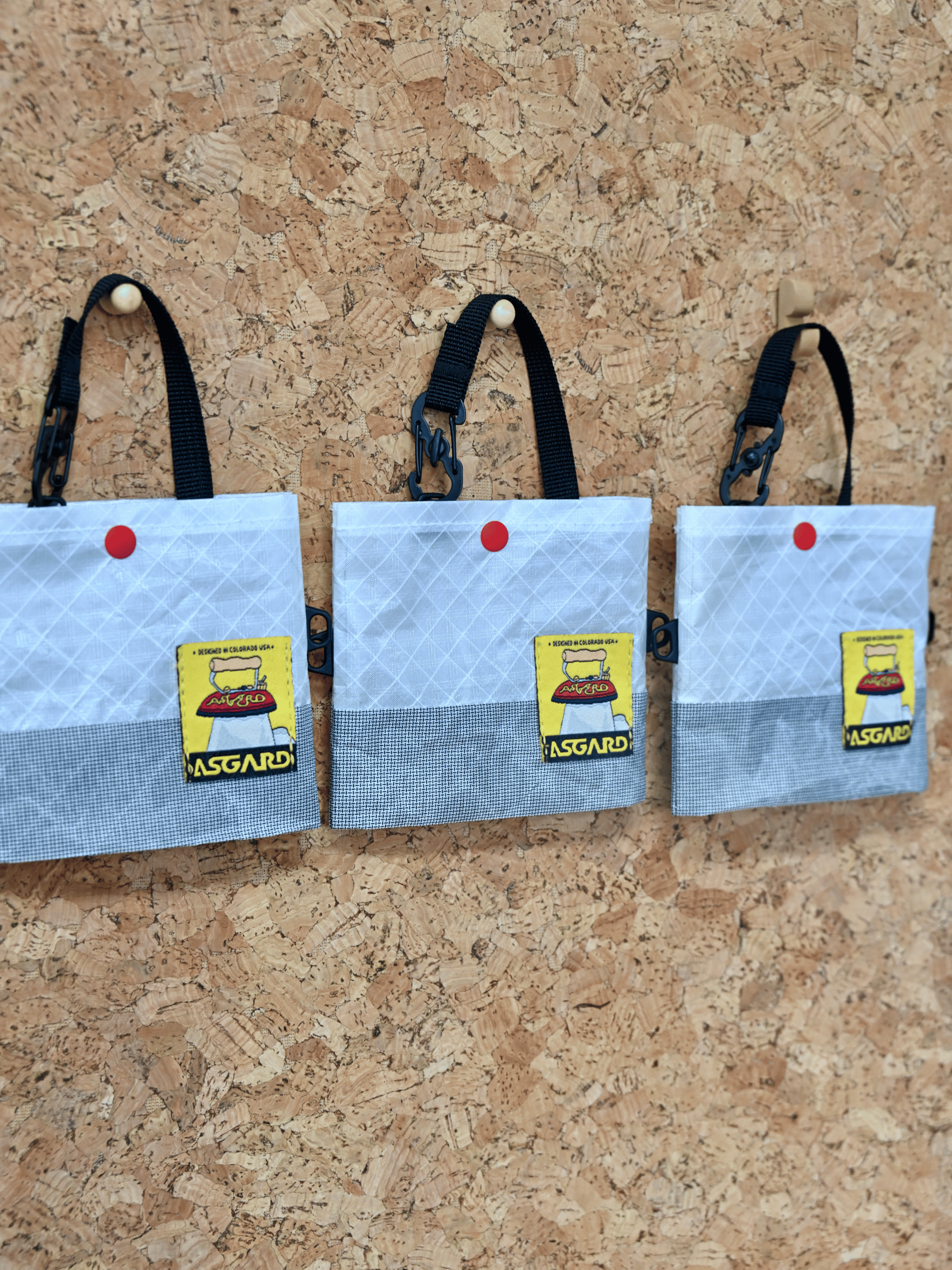 Three light gray Asgard Mini Tote Bags hanging by black handles against a cork wall.