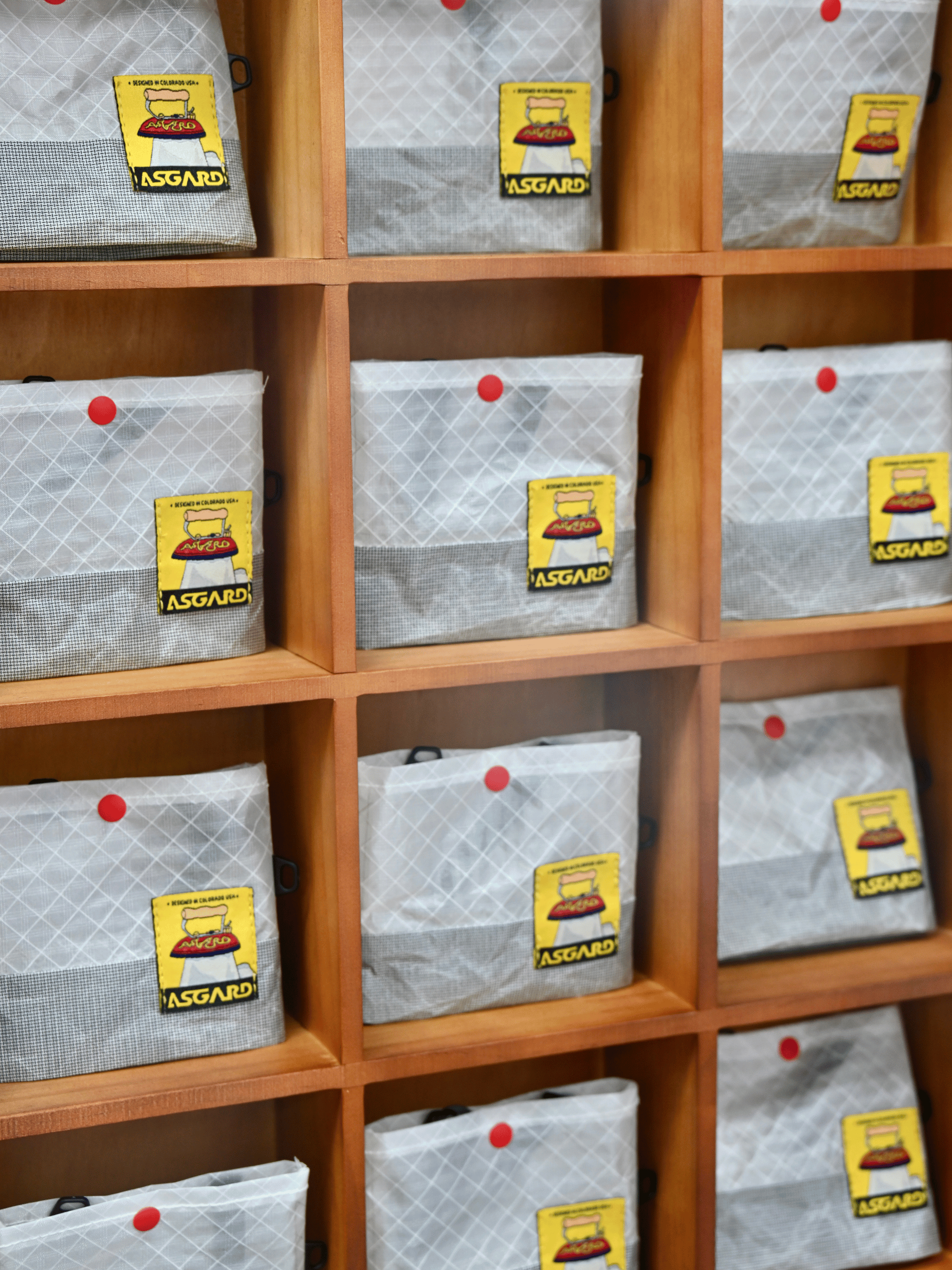 Light gray Asgard Mini Tote Bags arranged in wooden cubbies, with red snap closures and yellow logo patches.