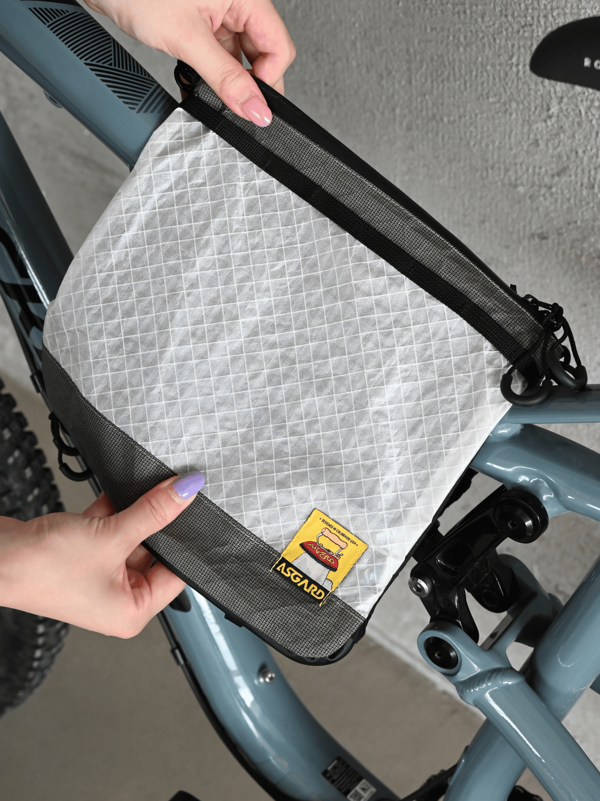 Hands holding the white Asgard Versa Bag 5L beside a bicycle frame, showing its top zipper and loop pulls.